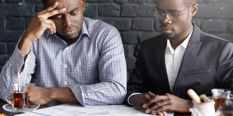 Candid shot of two handsome African-American business partners having frustrated looks, focused on working through papers, reviewing finances, trying to cope with financial stress in their company